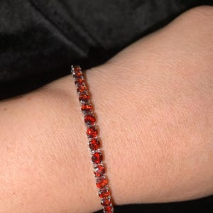 May include: A silver bracelet with a line of oval-cut, vibrant red gemstones. The bracelet is worn on a person's wrist, with the gemstones catching the light. The background is a dark, solid color.