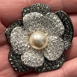 May include: A flower-shaped brooch with a pearl center and petals adorned with sparkling crystals. The outer petals are dark gray, while the inner petals are silver. This elegant accessory adds a touch of glamour.