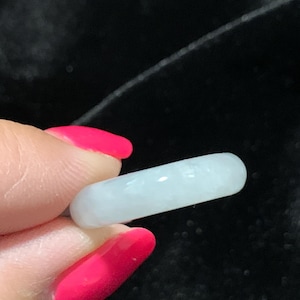 May include: A light blue, cylindrical jade ring. The ring is smooth and polished, with a slightly translucent appearance. The jade has a subtle, natural variation in color. The ring is held against a dark background.