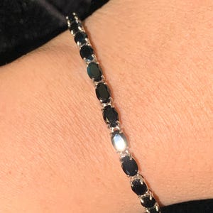 May include: A silver bracelet featuring a series of oval-cut, dark gemstones. The gemstones are set closely together, creating a continuous line of black against the silver setting. The bracelet is shown on a person's wrist.