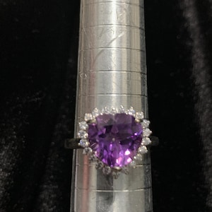 May include: A silver ring featuring a large, heart-shaped, purple gemstone. The gemstone is surrounded by smaller, clear stones. The ring is displayed on a silver ring sizer against a dark background.