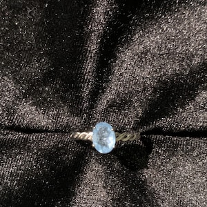 May include: A ring featuring a central oval-cut light blue gemstone held by four prongs. The band has a twisted rope-like texture, crafted from a metallic material that reflects light, creating a distinct contrast against the dark surface.