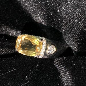 May include: A silver-toned ring featuring a large, rectangular, yellow gemstone at the center, surrounded by a halo of small clear stones. The band includes a small heart-shaped accent set with clear stones on the side of the metal shank.