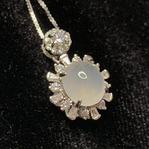 May include: A silver necklace with a pendant featuring a large, round, pale-colored stone surrounded by smaller, clear stones. The pendant is attached to a delicate silver chain. The background is a dark, textured fabric.