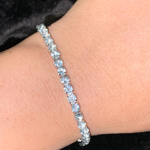 May include: A delicate bracelet featuring a series of light blue gemstones. The gemstones are set closely together, creating a continuous line of sparkle. The bracelet is a classic piece of jewelry.