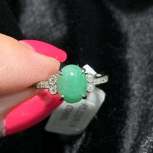 May include: Oval-shaped, light green gemstone ring with a silver band. The ring features small, clear accent stones and is held by a hand with pink nail polish. A white tag with text is visible.