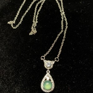 May include: A silver chain necklace with a teardrop-shaped pendant. The pendant features a green gemstone surrounded by clear rhinestones.