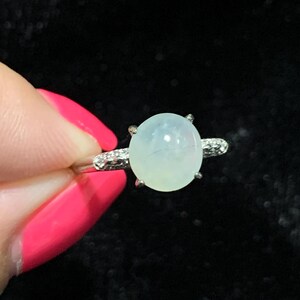 May include: A silver ring featuring a large, oval, pale green gemstone. The band is adorned with small, clear stones on either side of the main stone. The ring is held against a dark background.