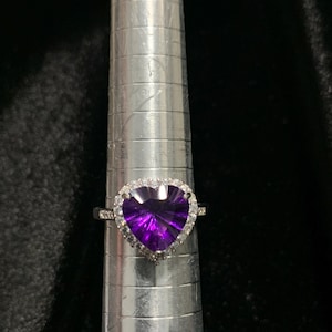 May include: A silver ring with a heart-shaped, deep purple gemstone. The gemstone is surrounded by a halo of smaller, clear stones. The ring is displayed on a silver ring sizer against a black background.