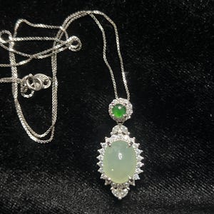 May include: A silver necklace with a pendant featuring a large, oval, pale green gemstone surrounded by small, clear stones. A smaller, round, green gemstone is above the oval stone. The necklace is on a black background.