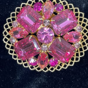 May include: A vintage pink rhinestone brooch with a gold tone metal setting. The brooch features a cluster of pink rhinestones in various shapes and sizes, surrounded by a delicate gold tone metal filigree design.