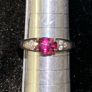 May include: A silver-toned ring featuring a central oval-cut vibrant pink gemstone flanked by three small clear round stones on each side. The ring is positioned on a metal ring mandrel marked with horizontal lines for size measurement.