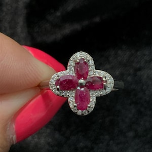 May include: A silver ring featuring a four-leaf clover design. Each leaf is a vibrant red gemstone, surrounded by a halo of smaller, clear stones. The ring is held against a dark background.