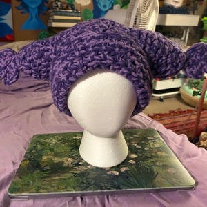 May include: A handmade, purple crochet hat with a distinctive design. The hat has extended, rounded elements on each side. The hat is displayed on a white mannequin head, resting on a laptop with a floral design.