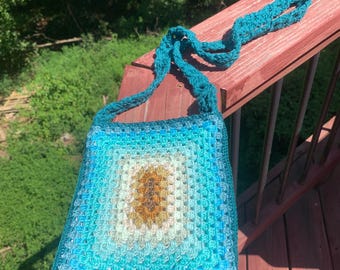 May include: A crocheted teal and blue ombre shoulder bag with a brown and white centre. The bag has a long teal crocheted strap.