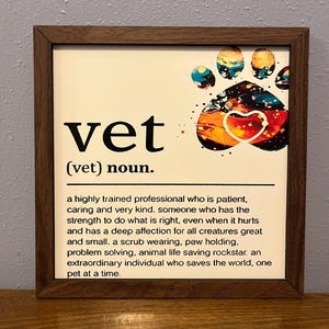 Può includere: Opera d'arte incorniciata con una cornice in legno scuro. L'opera presenta la parola "vet" in grandi lettere nere, con una definizione sottostante. Un colorato disegno di impronta di zampa e cuore si trova nell'angolo in alto a destra.