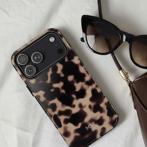 Puede incluir: Accesorios variados sobre fondo blanco: funda de teléfono con estampado de carey con iniciales "MSP", gafas de sol negras de ojo de gato, bolso de cuero marrón, pintalabios y anillo de madera.