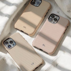 Puede incluir: Tres fundas de teléfono a rayas beige y marrón con iniciales personalizadas. Las fundas están dispuestas sobre una tela blanca con estampado floral. Las iniciales son "ML", "RL" y "GF".