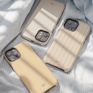 Puede incluir: Tres fundas para teléfono con patrones de rayas verticales en crema, amarillo y colores pastel. Cada funda tiene un monograma inicial diferente. Las fundas están sobre una tela a cuadros azul y blanco.