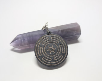 Puede incluir: Un colgante de madera negro con una estrella de cinco puntas tallada y un patrón circular. El colgante está colgado de una cadena plateada y descansa sobre un cristal morado.