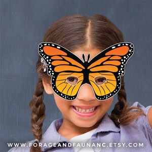 May include: A child wearing a paper butterfly mask with orange and black wings. The mask has eye holes and is held in place with a string. The child is smiling and has brown hair.