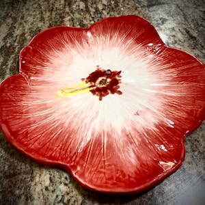May include: A red and white ceramic plate shaped like a hibiscus flower. The plate has a raised center and a textured surface.