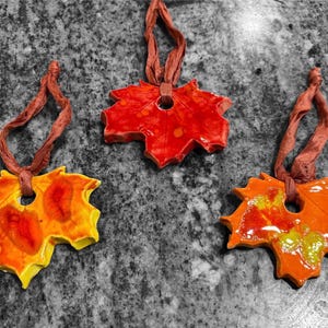 May include: Three ceramic leaf ornaments in shades of red, orange, and yellow, each with a brown ribbon for hanging. The leaves have a glossy finish and are arranged on a gray and black speckled surface. The ornaments are likely for fall decor.