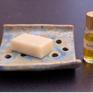 May include: A ceramic soap dish with a bar of soap and a small bottle of body oil. The soap dish is blue and brown with a dragonfly design. The body oil bottle has a wooden cap and a label that reads "Body Oil".