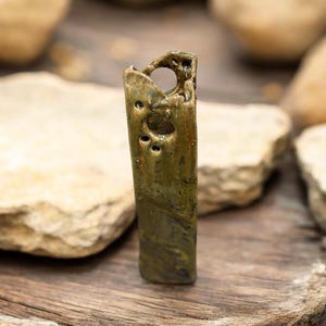 May include: A handcrafted, rectangular pendant with a weathered, metallic finish. The pendant features a hole at the top and several smaller holes along its length. The color is a mix of olive green and gold, with a textured surface.