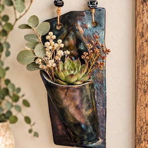 May include: A wall-mounted ceramic planter with a dark, iridescent glaze featuring metallic blue, purple, and bronze tones. The planter holds a green succulent and dried foliage, suspended by a jute rope with two black and white patterned beads.