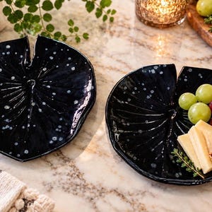 May include: Two black ceramic plates shaped like lily pads with a glossy finish and white speckled details. One plate is empty, while the other holds green grapes, red grapes, cheese slices, and crackers on a marble surface.