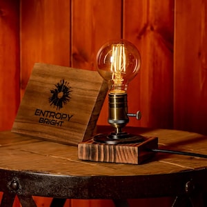May include: A rustic wooden table lamp with a vintage Edison bulb. The lamp is on a wooden base and has a black cord. The lamp is sitting on a wooden table with a sign that says "Entropy Bright".