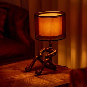 Puede incluir: Una lámpara de mesa única con diseño steampunk. La lámpara presenta una pantalla cilíndrica con un brillo cálido anaranjado, sostenida por una base hecha de tuberías metálicas interconectadas. La lámpara está sobre una mesa de madera.