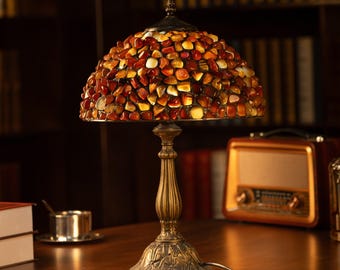 May include: A decorative table lamp with a domed shade composed of amber, brown, and cream-colored stones. The lamp has a bronze-colored base with a detailed floral design. The lamp is on a wooden surface.