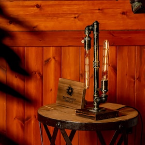 May include: A steampunk-style table lamp with two Edison bulbs. The lamp is made of black metal pipes and has a wooden base. The lamp is sitting on a wooden table with a metal base.