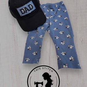 Op de afbeelding: Een zwarte baseballcap met het woord "DAD" op een blauwe patch, gecombineerd met een blauwe broek met bloemenprint. De broek heeft een wit en geel bloemenpatroon. De items liggen op een licht houten oppervlak.