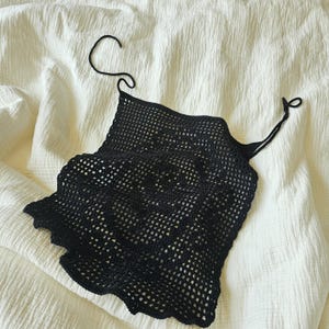 May include: A black crochet top with a square neckline and thin straps. The top has a scalloped edge and a net-like pattern. The garment is displayed on a white, textured surface.