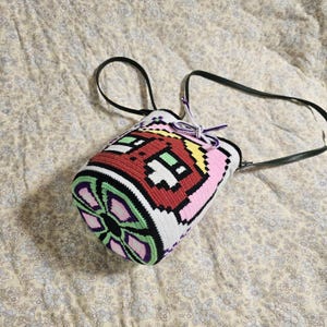 May include: A vibrant, hand-crocheted bag with a pixelated design. The bag showcases a red, pink, green, yellow, and white pattern, with a black strap and drawstring closure. The bag is cylindrical and appears to be made of yarn.