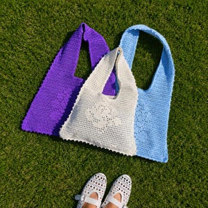 Può includere: Tre borse tote all'uncinetto nei colori viola, crema e azzurro, ognuna caratterizzata da un distinto motivo floreale integrato nel design a rete. Le borse hanno un unico ampio tracolla e un corpo quadrato con una struttura a trama aperta e testurizzata.