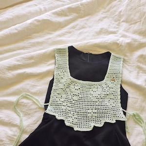 Peut inclure: Un haut en crochet vert clair avec un col carré et des bords festonnés, porté sur une robe noire. Le motif ajouré est visible. Le haut a des liens vert clair. Un petit bouton doré est sur le dessus.