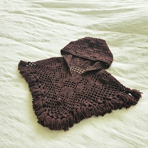 May include: A dark brown crochet poncho with a matching hood. The poncho features a fringed hem and a detailed openwork design. The hood is also crocheted with a similar pattern. The garment is displayed on a white surface.