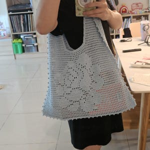 May include: A light blue crochet tote bag featuring an open-work mesh pattern and a central floral motif. The bag has a wide shoulder strap and a decorative border of small, evenly spaced crochet bobbles along the side edges of the fabric.