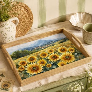May include: A rectangular wooden serving tray featuring a printed illustration of a vibrant sunflower field under a blue sky with distant mountains and a small village. The tray has integrated cutout handles on the sides and a light wood finish.