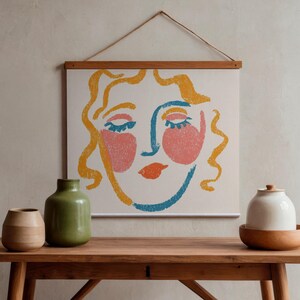 Könnte beinhalten: Ein Wandbehang mit einer stilisierten Strichzeichnung eines Gesichts mit gelbem Haar, blauen Gesichtskonturen, rosa Wangen und orangefarbenen Lippen. Das Kunstwerk wird von einem Holzrahmen mit einer Schnur gehalten und steht auf einem Holztisch.