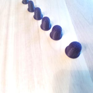 Könnte beinhalten: Sechs dunkelviolette Fingerhüte mit einer genoppten Oberfläche sind diagonal auf einer hellen Holzmaserung angeordnet. Die Fingerhüte bestehen aus einem festen Material und haben eine klassische Form.