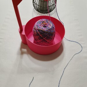 Könnte beinhalten: Ein rosa Garnhalter mit einer Schüssel, die eine Kugel mehrfarbigen Garns enthält, und einem Haken, um das Garn zu halten.
