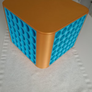May include: A cube-shaped storage container with a flat, orange top and sides. The sides feature a blue, textured pattern of oval indentations. The container is sitting on a white surface.