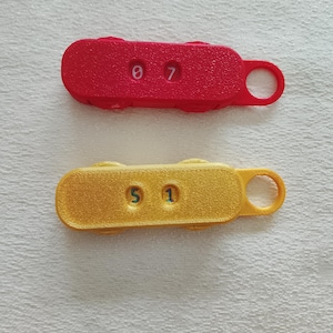 Könnte beinhalten: Zwei rechteckige Schlüsselanhänger aus Kunststoff. Einer ist rot mit den Zahlen 0 und 7. Der andere ist gelb mit den Zahlen 5 und 1. Jeder Schlüsselanhänger hat an einem Ende eine kleine Schlaufe.