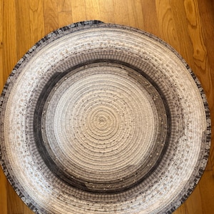 May include: Circular, neutral-toned rug with concentric patterns. The rug features a spiral design in shades of white, gray, and black. The edges have a patterned border. The rug is made of a woven material.