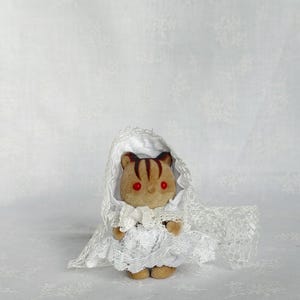 May include: A small, flocked animal figurine with brown fur, dark brown stripes on its head, and bright red eyes. The figure is dressed in a white lace bonnet and a matching white lace dress with a ruffled skirt and a small bow at the neck.